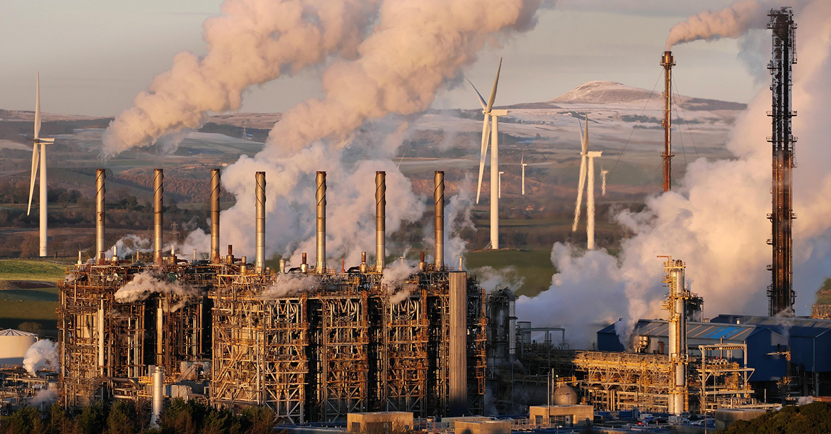 Industrial petrochemical plant with steam rising from tall stacks at sunrise, wind turbines and hills in the background